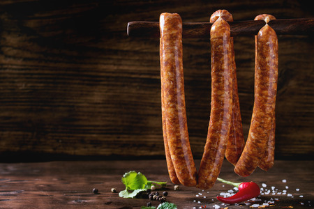 Fresh raw beef sausages with spices for BBQ hanging on stick over dark wooden background with salt, herbs and pepper. With space for textの写真素材
