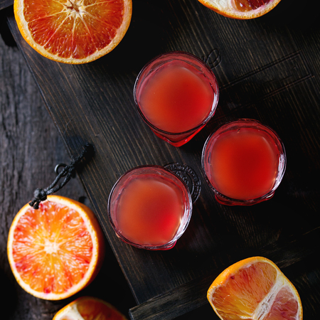 Shorts of alcohol cocktail with Sliced Sicilian Blood oranges and fresh red orange juice, served on black wooden chest over old wooden table. Dark rustic style. Top view. Square image with selective focusの写真素材