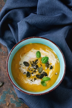 Turquoise ceramic bowl of pumpkin and sweet potato cream soup with fresh basil, cream and seeds, served on textile napkin over old wood textured background. Top viewの写真素材
