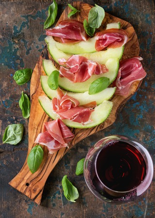 Sliced melon with ham and basil leaves, served on olive wood chopping board with glass of red wine over old wooden textured background. Top viewの写真素材