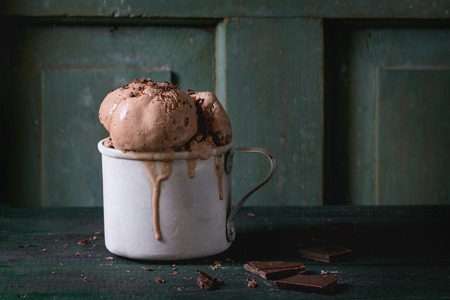 Frozen vintage aluminum mug with melting chocolate ice cream balls, served with chopped dark chocolate on old wooden table. Dark rustic style. Space for textの写真素材