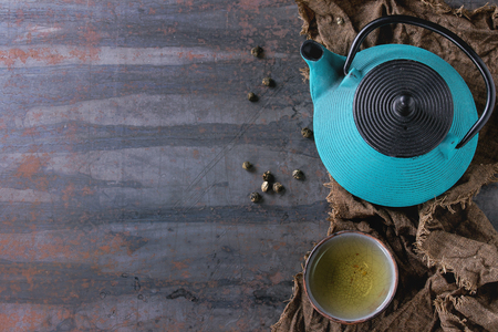 Handmade ceramic cup of hot green tea, served with blue iron teapot on wet sackcloth over old dark textured background. Asian style. Top view with copy sapceの写真素材
