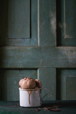Frozen vintage aluminum mug with melting chocolate ice cream balls, served with chopped dark chocolate on old wooden table. Dark rustic style. Space for textの写真素材