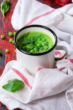 White vintage mug with pea cream soup with sliced basil, served with red salt and pepper shakers on white kitchen towel over red and black wooden background.の写真素材