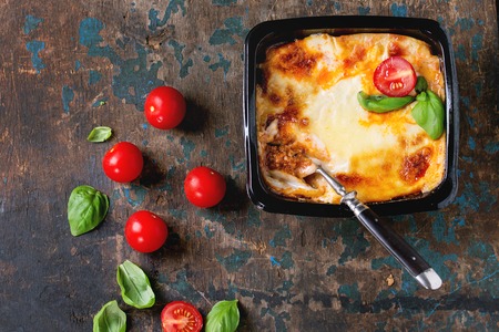Cooked meat lasagna in black plastic box, served with vintage fork,  fresh cherry tomatoes and basil leaves over old dark wooden textured background. Market semifinished catering theme. Top viewの写真素材