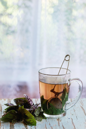 Glass cup of hot herbal tea with bunch of fresh violet basil, served with vintage tea-strainer on old wooden stool with window at background. Rustic style, natural day light.の写真素材