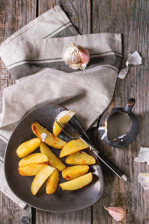 Fried country potatoes on brown ceramic plate, served with homemade aioli sauce in jug, garlic and vintage fork on gray linen napkin over wooden background. Rustic style. Top viewの写真素材
