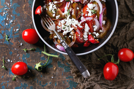 Black bowl with sliced different tomatoes, red onion, balsamic sause, thyme and feta cheese salad, served with fork and cherry tomato on sackcloth over old dark wood textured background. Top viewの写真素材