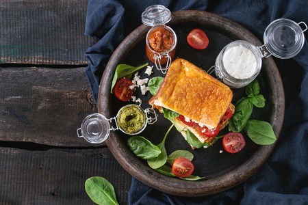 Low-carb gluten free Cloud bread veggie sandwich with spinach, avocado, feta cheese, tomatoes and pesto sauce, served on clay tray with jars of ingredients over old wood background. Top viewの写真素材