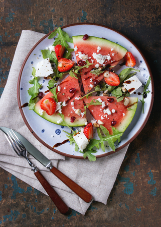 Blue spotted plate with watermelon and strawberry fruit salad with feta cheese, arugula, nuts and balsamic sauce, served with fork, knife and napkin over old dark wood background. Top view.の写真素材