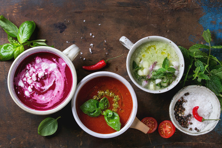 Variety of cold vegetarian soups gazpacho cucumber with mint, beetroot with feta chhese, tomato with green pesto, served with seasoning in cups over wood background. Top view copy spaceの写真素材