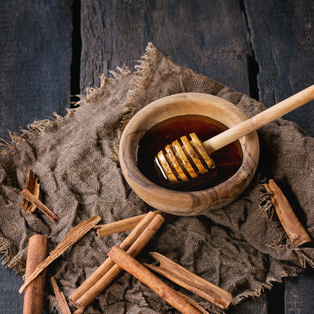 Honey in olive wood bowl with honey-dipper and cinnamon sticks on sackcloth rag over old black wooden background. Dark rustic style. Square imageの写真素材