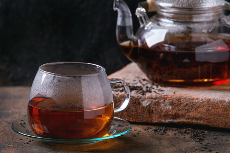 Glass cup with saucer and teapot with hot black tea, standing on clay board over old wooden background.の写真素材