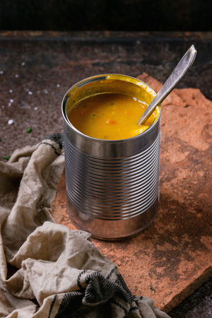 Tin can of carrot soup with spoon, standing on kitchen towel  and clay board over old rusty iron background. Dark rustic styleの写真素材