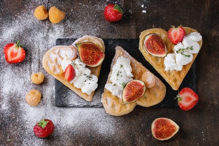 Wafers as heart shape with yellow raspberries, strawberries, sliced figs, ricotta cheese and sugar powder on black slate board over dark wooden background. Top view with space for textの写真素材