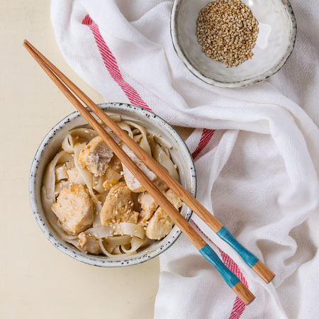 Ceramic bowl of udon noodles with chicken pieces and sesame seeds, served with wood chopsticks on textile towel over white wooden background. Top view. Asian style.の写真素材