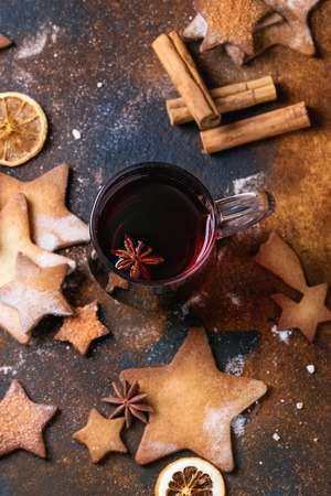 Glass mug of hot red mulled wine spices, sugar shortbread cookies star shape, anise and cinnamon powder over dark textured background. Christmas drink treat theme. Overhead viewの写真素材