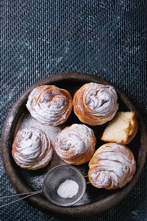 Modern pastries cruffins, whole and slice, like croissant and muffin with sugar powder, served in clay tray with vintage sieve over dark texture background. Top view. Space for textの写真素材