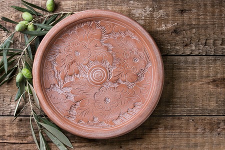 Empty clay ceramic ornate plate and young olives branch over old wood background. Overhead view with space for textの写真素材