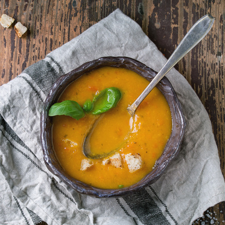 Ceramic bowl of carrot soup, served with fresh basil, croutons, kitchen towel and spoon on old wooden background. Dark rustic style, overhead view. Square imageの写真素材