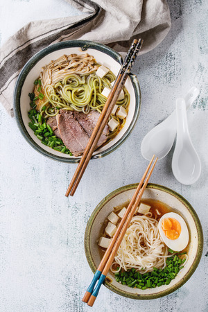 Two bowl of different asian style soups with green tea soba noodles, egg, mushrooms, beef, onion and tofu cheese, served with chopsticks and spoon over white wooden background. Top viewの写真素材