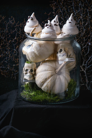 Halloween sweet dessert table decor glass jar with white meringue ghosts with chocolate eyes, decor skulls, moss and pumpkin over black background.の写真素材