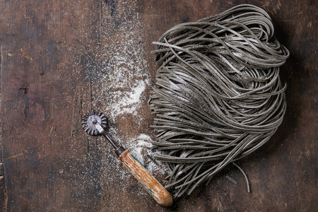 Raw uncooked black cuttlefish ink spaghetti pasta with flour and dough disk cutter over dark wooden background. Top view with space for text.の写真素材