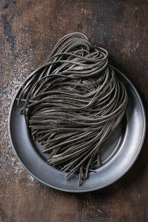 Raw uncooked black cuttlefish ink spaghetti pasta with flour on vintage metal plate over dark wooden background. Top view with space for text.の写真素材