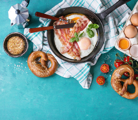 Breakfast with fried eggs and bacon in cast-iron pan, broken egg, tomatoes, mustard, pretzels and basil on white kitchen towel over turquoise wooden background. Top view with copy spaceの写真素材