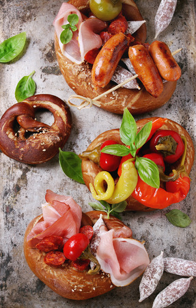 Variety of meat snacks fried sausages, wienerwurst, ham, marinated chili peppers served in salted pretzels with fresh basil over old metal background. Top viewの写真素材