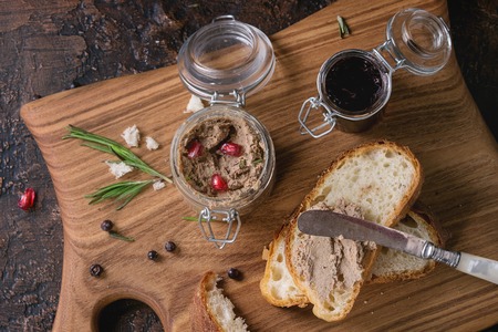 Glass jars of chicken liver pate with blackcurrant jam, pomegranate grain and sliced bread, served with vintage knife on wooden chopping board over dark texture background. Top viewの写真素材