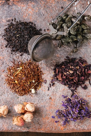 Variety of black, green, rooibos, herbal dry tea leaves and rose buds with vintage strainer on terracotta board over old dark wooden background. Top view, close upの写真素材