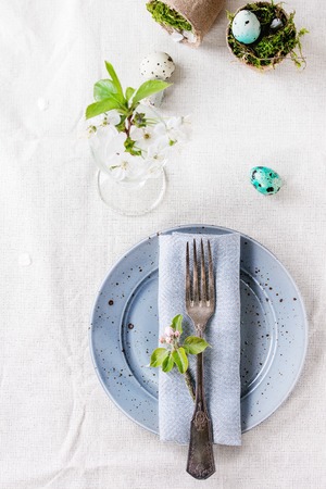 Table setting decor colorful Easter quail eggs with spring cherry flowers, moss in garden pots, empty plates, vintage cutlery over white tablecloth. Top view with copy space.の写真素材