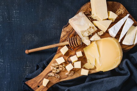 Cheese plate. Assortment of cheese with walnuts, honey from honey dipper on olive wood serving board with textile over dark blue canvas as background. Top view with space.の写真素材