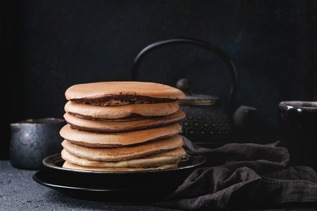 Stack of homemade american ombre chocolate pancakes served on black plate with jug of cream and teapot over black stone texture background.の写真素材