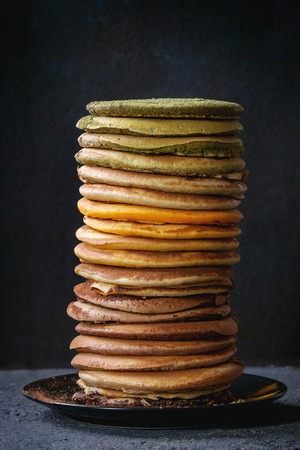 High stack of colorful homemade american ombre chocolate, green tea matcha and turmeric pancakes on plate over black stone texture background.の写真素材