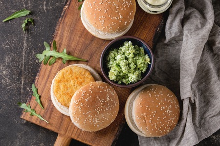 Ingredients for making vegan burger. Veggie cheese and onion cutlets, yogurt sauce, hamburger buns, herbs and avocado salad, served on wooden board with textile on dark background. Top viewの写真素材