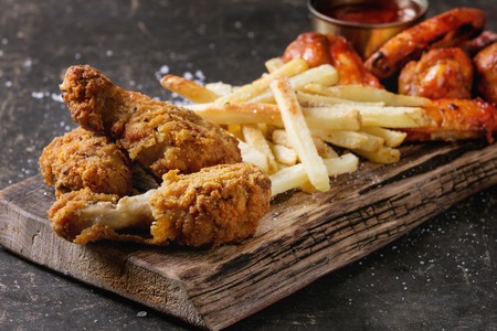 Fast food fried crispy chicken legs and french fries potatoes with salt served on wooden serving board over dark texture background. Space for text. Close upの写真素材