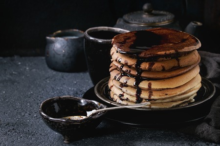 Stack of homemade american ombre chocolate pancakes with carob honey sauce served on black plate with jug of cream and teapot over black stone texture background.の写真素材