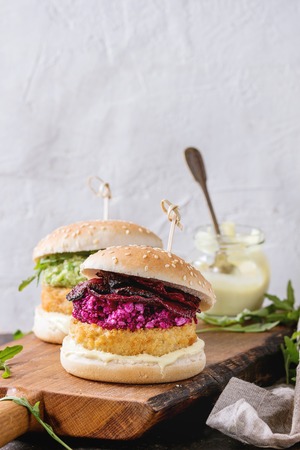 Two vegetarian hamburgers with onion and cheese cutlets, avocado salad, arugula, fried beetroot and yogurt sauce on wooden serving board over texture background. Healthy fast food.の写真素材