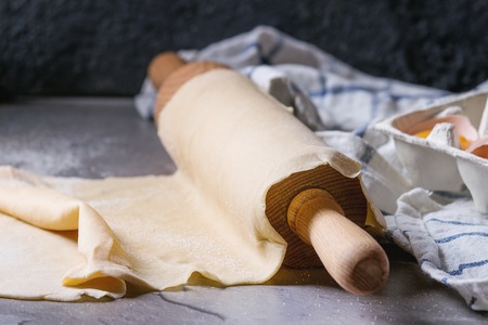 Fresh homemade rolled dough for pasta tagliatelle on wood rolling pin with egg yolk and kitchen towel over dark gray table background.の写真素材