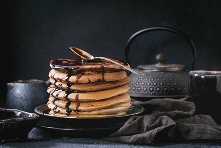 Stack of homemade american ombre chocolate pancakes with carob honey sauce served on black plate with jug of cream and teapot over black stone texture background.の写真素材