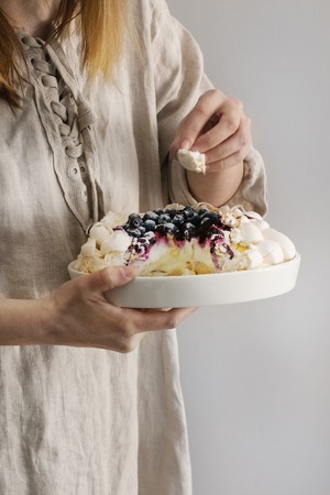 Woman tasting cake. Homemade meringue cake Pavlova with whipped cream, sugar powder, fresh blueberries and blueberry sauce in female hands over gray wall as background, day light.の写真素材