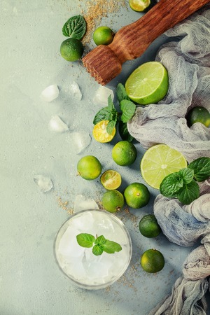 Ingredients for mojito cocktail, whole, sliced lime and mini limes, mint leaves, brown crystal sugar over gray stone texture background, gauze textile, bar muddler, glass of ice. Top view, spaceの写真素材