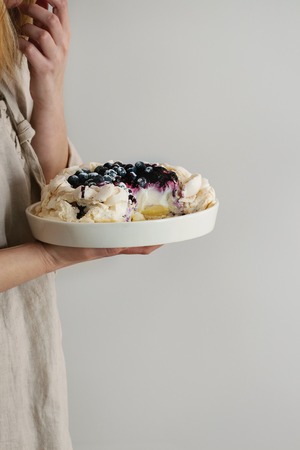 Woman tasting cake. Homemade meringue cake Pavlova with whipped cream, sugar powder, fresh blueberries and blueberry sauce in female hands over gray wall as background, day light.の写真素材
