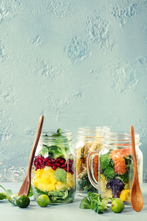 Variety of salads in mason jars. Fruit salad mango, pomegranate, greens, vegetables pasta, carrots, cauliflower, salmon. Standing with mini limes and wood spoon over blue background. Food to go.の写真素材