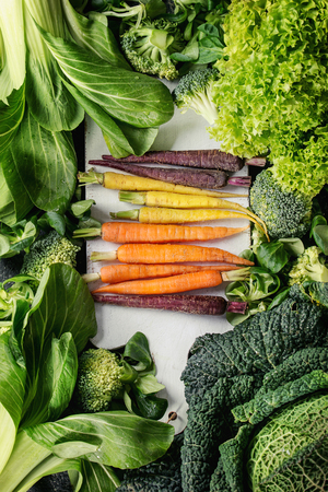 Variety of raw green vegetables salads, lettuce, bok choy, corn, broccoli, savoy cabbage, colorful young carrots on white chopping board. Food background. Top viewの写真素材