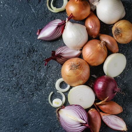 Variety of whole and sliced red, white, yellow and shallot onions over dark stone texture background. Top view, space for text. Square imageの写真素材