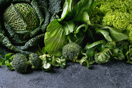 Variety of raw green vegetables salads, lettuce, bok choy, corn, broccoli, savoy cabbage as frame over black stone texture background. Space for text