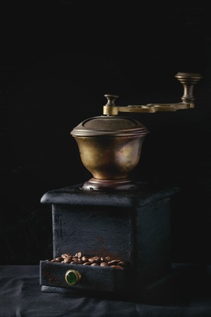 Old vintage grinder with roasted coffee beans and grind coffee in tin jar with scoop over black table with black wooden background. Dark still lifeの写真素材